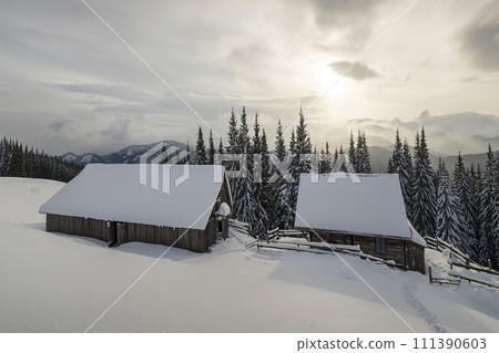 Winter mountain landscape. Old wooden houses on snowy clearing on background of mountain ridge, spruce forest and cloudy sky. Happy New Year and Merry Christmas card. 111390603