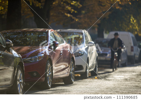 Long row of different shiny cars and vans parked along empty roadside on sunny autumn day on blurred green golden foliage bokeh background. Modern city lifestyle, vehicles parking problem concept. Long row of different shiny cars and vans parked along empty roadside on sunny autumn day on blurred green golden foliage bokeh background. Modern city lifestyle, vehicles parking problem concept. 111390685