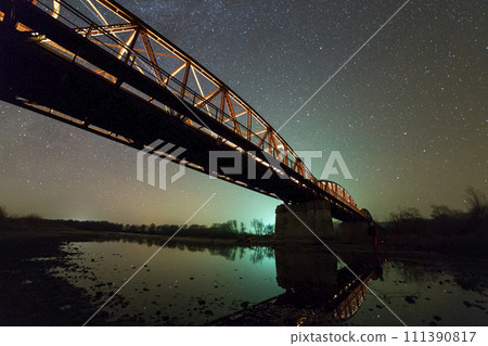 Illuminated metal bridge on concrete supports reflected in water on dark starry sky with Milky Way constellation background. Night photography concept. 111390817