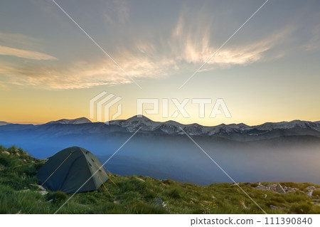 Summer camping in mountains at dawn. Tourist tent on round grassy hill on distant misty blue mountains range under pink sky before sunrise or sunset. Tourism, hiking and beauty of nature concept. Summer camping in mountains at dawn. Tourist tent on round grassy hill on distant misty blue mountains range under pink sky before sunrise or sunset. Tourism, hiking and beauty of nature concept. 111390840