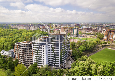 Apartment or office tall building unfinished under construction among green tree tops. Tower cranes on bright blue sky copy space background. Apartment or office tall building unfinished under construction among green tree tops. Tower cranes on bright blue sky copy space background. 111390873