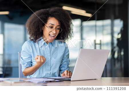 A woman is happily using her laptop computer while sitting at a desk in a room with fun decor and a smile on her face. 111391208