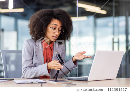 A woman is seated at a desk with a laptop, microphone, and office supplies. A woman is seated at a desk with a laptop, microphone, and office supplies. 111391217