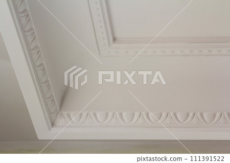 Ornamental white molding decor on ceiling of white room close-up detail. Interior renovation and construction concept. Ornamental white molding decor on ceiling of white room close-up detail. Interior renovation and construction concept. 111391522