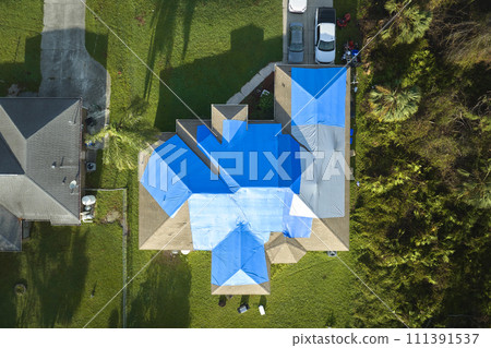 Aerial view of damaged in hurricane Ian house roof covered with blue protective tarp against rain water leaking until replacement of asphalt shingles 111391537