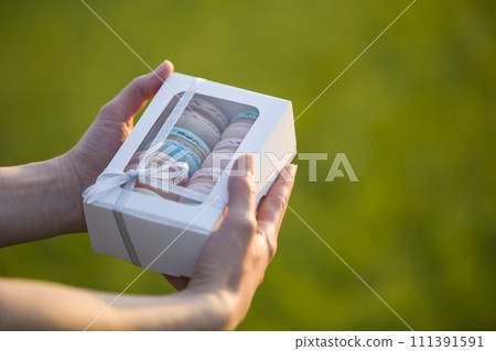 Female hands holding cardboard gift box with colorful pink blue handmade macaron cookies on green blurred copy space background. 111391591