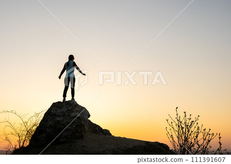 Silhouette of a woman hiker standing alone on big stone at sunset in mountains. Female tourist on high rock in evening nature. Tourism, traveling and healthy lifestyle concept. 111391607