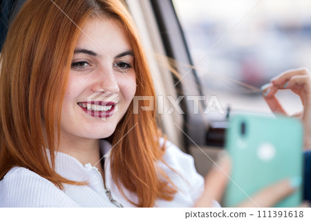 Young redhead woman driver taking selfies with her mobile phone sitting behind the wheel of the car in rush hour traffic jam. 111391618