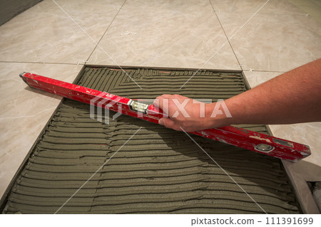 Ceramic tiles and tools for tiler. Worker hand installing floor tiles. Home improvement, renovation - ceramic tile floor adhesive, mortar, level. Ceramic tiles and tools for tiler. Worker hand installing floor tiles. Home improvement, renovation - ceramic tile floor adhesive, mortar, level. 111391699