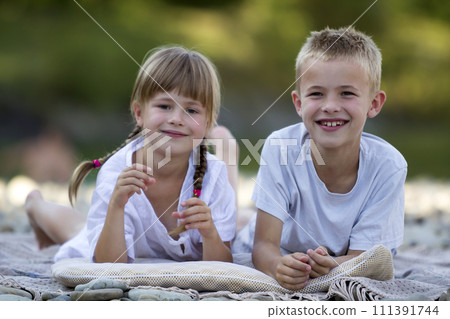 Two young happy cute blond children, boy and girl, brother and sister laying on pebbled beach on blurred bright sunny summer day background. Siblings friendship, leisure and perfect holiday concept. 111391744