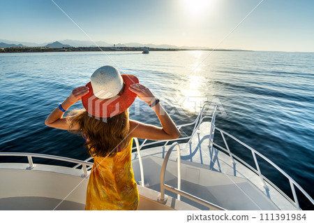 Young woman with long hair wearing yellow dress and straw hat standing on white yacht deck enjoying view of blue sea water. Young woman with long hair wearing yellow dress and straw hat standing on white yacht deck enjoying view of blue sea water. 111391984