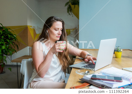 Focused young businesswoman or student looking at laptop, having online meeting or educational course, drinking tea in cafe, coworking. Remote work, social networks, blogging. Freelancer. 111392264