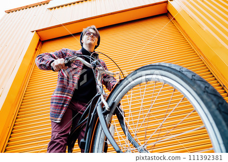 Bottom view Young stylish hipster man standing with a cruiser city Bicycle near orange Wall. Student Resting after ride bicycle. Eco friendly transportation for carbon footprint. Sustainable lifestyle Bottom view Young stylish hipster man standing with a cruiser city Bicycle near orange Wall. Student Resting after ride bicycle. Eco friendly transportation for carbon footprint. Sustainable lifestyle 111392381