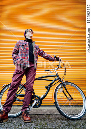 Young stylish hipster man standing with a cruiser city Bicycle near orange Wall. Student Resting after ride bicycle. Eco friendly transportation for carbon footprint. Sustainable lifestyle. Vertical 111392382