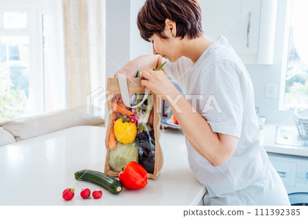 Woman removes fresh organic vegetables from reusable eco sackcloth fabric bag on the white kitchen table. Home food delivery concept. Local farmer healthy food. Eco-friendly, Sustainable lifestyle. Woman removes fresh organic vegetables from reusable eco sackcloth fabric bag on the white kitchen table. Home food delivery concept. Local farmer healthy food. Eco-friendly, Sustainable lifestyle. 111392385