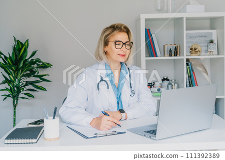 Confident mature caucasian senior female doctor in white coat using laptop for telemedicine with patients at hospital. GP make online video call consult patient. Online remote medical appointment. 111392389