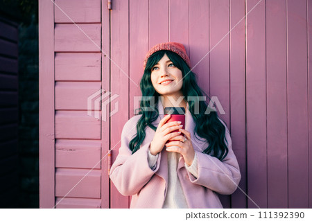 Relaxed stylish young smiling hipster woman wearing color hair, pink coat, hat holding reusable coffee cup leaning on beach hut and enjoying moment. Simple pleasures and personal fulfillment. 111392390