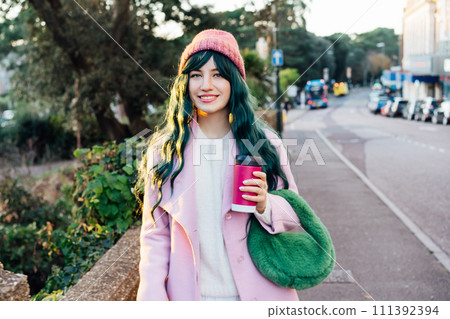 Stylish young smiling hipster woman with color hair walking on street in pink outfit with reusable coffee cup wearing coat, knitted hat, fur bag, happy mood, seasonal fashion, Barbiecore style trend Stylish young smiling hipster woman with color hair walking on street in pink outfit with reusable coffee cup wearing coat, knitted hat, fur bag, happy mood, seasonal fashion, Barbiecore style trend 111392394