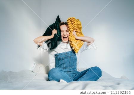 frustrated woman covers ears with pillows, screaming feels furious exhausted exasperated sitting on bed. mad irritated millennial suffers from head ache lack of self-control. Noise and stress concept. 111392395