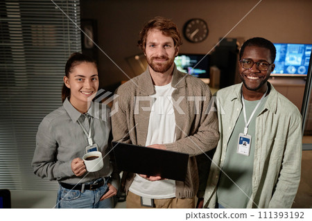 Waist up portrait of three young people holding laptop and looking at camera while standing in IT office at night Waist up portrait of three young people holding laptop and looking at camera while standing in IT office at night 111393192