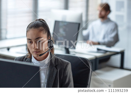 Portrait of young woman wearing headset with microphone and using computer while working in customer support office copy space Portrait of young woman wearing headset with microphone and using computer while working in customer support office copy space 111393239