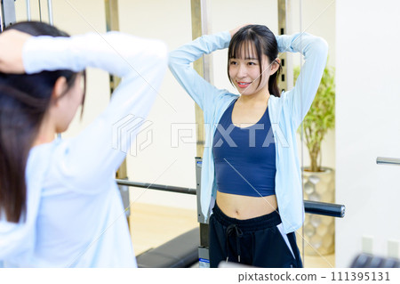 Young woman checking her figure in the gym mirror and getting ready. Private gym, muscle training girl, tying her hair. Young woman checking her figure in the gym mirror and getting ready. Private gym, muscle training girl, tying her hair. 111395131