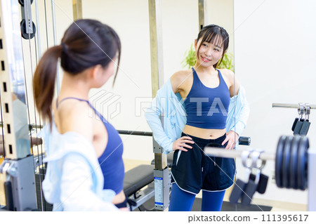 一位年輕女子在私人健身房照鏡子檢查自己的身材。私人健身房，肌肉訓練的女孩。 111395617