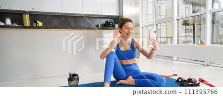 Image of beautiful, atheltic girl sits on yoga mat at home with smartphone, takes selfie and workout, does fitness exercises 111395766