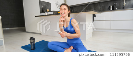 Portrait of sportswoman using sport app while doing workout, yoga exercises at home, sits on mat with water bottle, smiling and looking happy 111395796