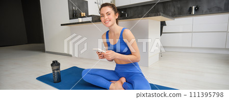 Sport and wellbeing. Young sportswoman does exercises at home, sits in yoga pose, holds smartphone, follows online training videos during her workout indoors 111395798