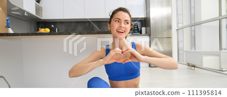 Portrait of beautiful, fit young woman in leggins and sportsbra, sits in lotus pose on yoga mat at home, shows heart sign and smiles at camera, workout, fitness instructor online video training 111395814