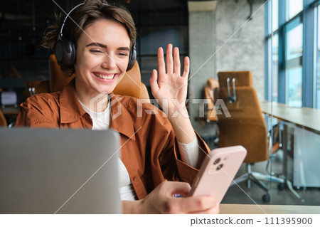 Portrait of working office woman sitting with laptop, joins video chat, records video for company social media in wireless headphones, waves hand at smartphone app 111395900