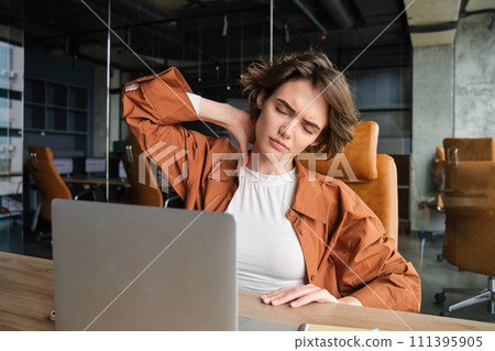 Portrait of woman feels pain in neck, sits in office with laptop, has tension in muscles after working all day 111395905