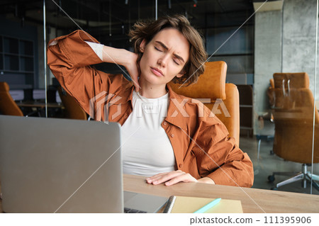 Image of woman with tired face, sits with laptop in office, feels tension in neck, pain in muscles after working all day at workplace 111395906