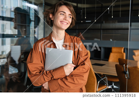 Portrait of young female manager, office worker with laptop, digital nomad programmer working at her workplace, standing with computer and smiling 111395918