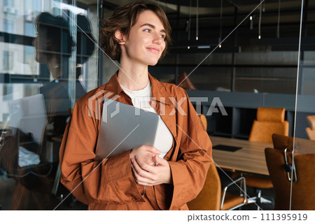 Portrait of young female manager, office worker with laptop, digital nomad programmer working at her workplace, standing with computer and smiling 111395919