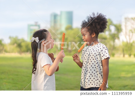 Happy cheerful white girls and black girl eat sausage together at outdoors park , Relationship little kids, Diverse ethnic concept. Happy cheerful white girls and black girl eat sausage together at outdoors park , Relationship little kids, Diverse ethnic concept. 111395959