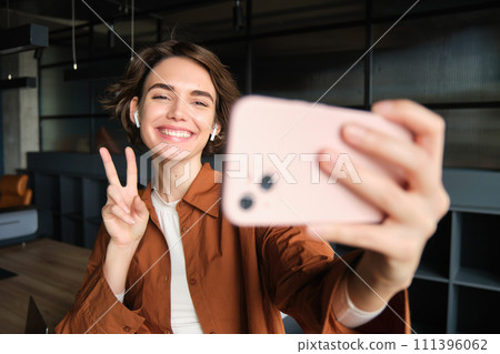 Portrait of beautiful smiling woman, taking selfie on smartphone, connects to online streaming, creating content for company, showing peace sign 111396062