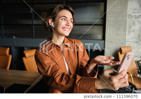 Portrait of woman in wireless headphones video chats, connects to a meeting on smartphone, has a live streaming on mobile phone, stands in an office Portrait of woman in wireless headphones video chats, connects to a meeting on smartphone, has a live streaming on mobile phone, stands in an office 111396070