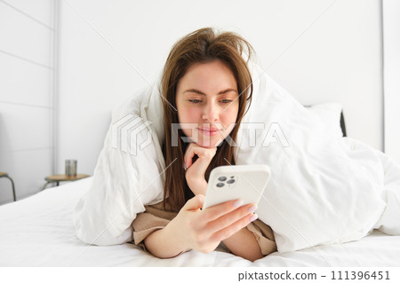Portrait of cute woman lying in bed and using smartphone, browsing online social media and resting in bedroom in morning Portrait of cute woman lying in bed and using smartphone, browsing online social media and resting in bedroom in morning 111396451