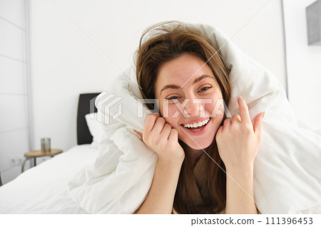 Portrait of beautiful brunette woman lying in bed, resting in her hotel room, covered in white blankets, laughing and smiling at camera Portrait of beautiful brunette woman lying in bed, resting in her hotel room, covered in white blankets, laughing and smiling at camera 111396453