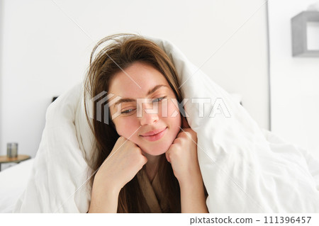 Cute girl with messy hair, lying in bed covered in white sheets duvet, smiling and laughing coquettish, spending time in her bedroom Cute girl with messy hair, lying in bed covered in white sheets duvet, smiling and laughing coquettish, spending time in her bedroom 111396457