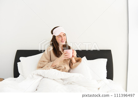 Portrait of beautiful woman in sleeping bag and pyjamas, drinking coffee in bed, enjoying her bright, relaxing morning Portrait of beautiful woman in sleeping bag and pyjamas, drinking coffee in bed, enjoying her bright, relaxing morning 111396497