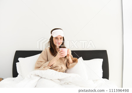 Gorgeous woman in pyjamas and sleeping mask, holding cup of coffee, drinking cappuccino in her bed and looking happy, resting in bedroom on weekend, having relaxing morning 111396498
