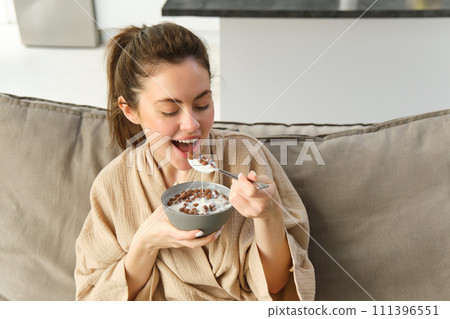 Portrait of gorgeous brunette woman sits on sofa, having her breakfast at home, smiling while eating cereals, holding bowl and spoon and laughing 111396551