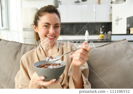 Relaxing morning at home. Young beautiful woman in bathrobe, eating cereals on sofa, having her breakfast in living room 111396552