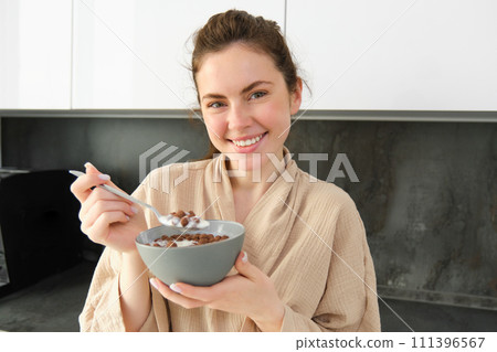 Gorgeous woman starts her day with cereals, eats breakfast and smiles, wears bathrobe, leans on kitchen worktop, starts her morning with quick meal 111396567