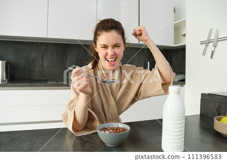 Portrait of enthusiastic young woman eating cereals with milk, looking excited and happy, sitting near kitchen worktop and having breakfast, raising hand up in triumph Portrait of enthusiastic young woman eating cereals with milk, looking excited and happy, sitting near kitchen worktop and having breakfast, raising hand up in triumph 111396583