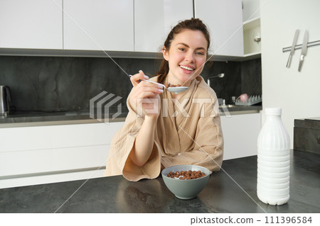 Portrait of happy young woman leans on kitchen worktop and eating cereals, has milk and bowl in front of her, having her breakfast, wearing bathrobe 111396584