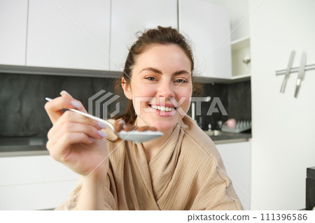 Portrait of smiling beautiful woman, feeding you spoon of cereals with milk, eating breakfast and looking happy, enjoying her morning, wearing cosy bathrobe 111396586